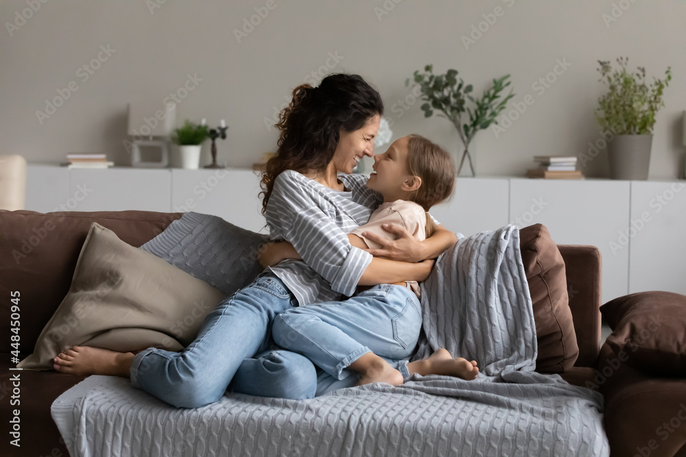 Happy mom and little daughter girl hugging, resting on couch, enjoying time together at home. Mother cuddling, embracing kid with love, tenderness. Family, being parent, motherhood concept