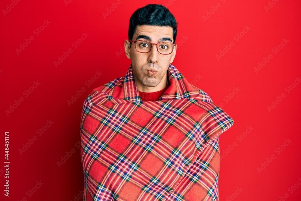 Handsome hispanic man wrapped in a red warm red blanket puffing cheeks with funny face. mouth inflated with air, catching air. Handsome hispanic man wrapped in a red warm red blanket puffing cheeks with funny face. mouth inflated with air, catching air.