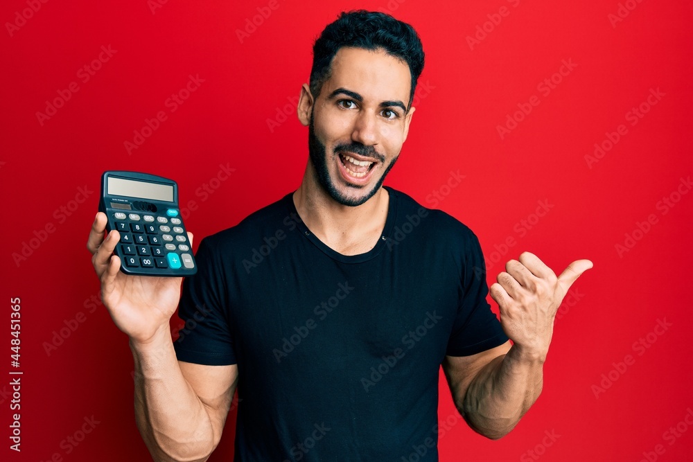Young hispanic man showing calculator device pointing thumb up to the side smiling happy with open mouth Young hispanic man showing calculator device pointing thumb up to the side smiling happy with open mouth