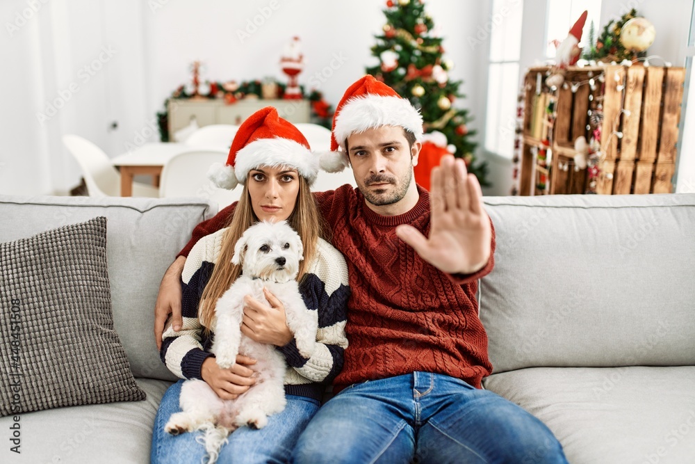 Obraz premium Young couple of wife and husband sitting on the sofa wearing christmas hat at home with open hand doing stop sign with serious and confident expression, defense gesture