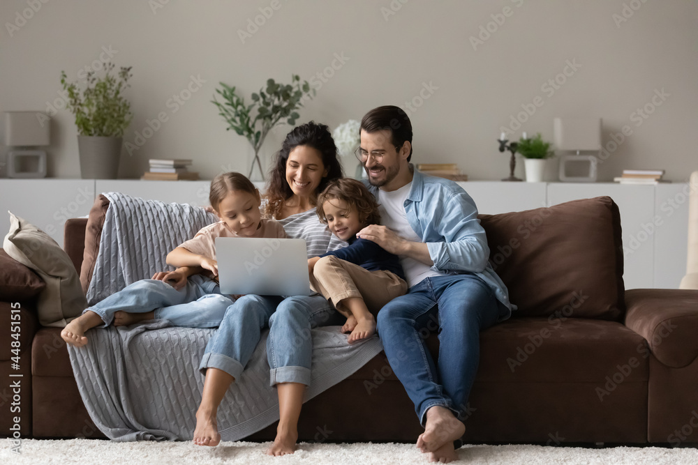 Happy millennial parents and gen Z kids relaxing on couch at home, using app on laptop, watching movie online, making video call, shopping on internet, playing virtual game on digital device