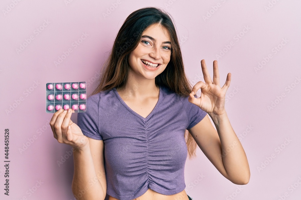 Young caucasian woman holding pills doing ok sign with fingers, smiling friendly gesturing excellent symbol Young caucasian woman holding pills doing ok sign with fingers, smiling friendly gesturing excellent symbol