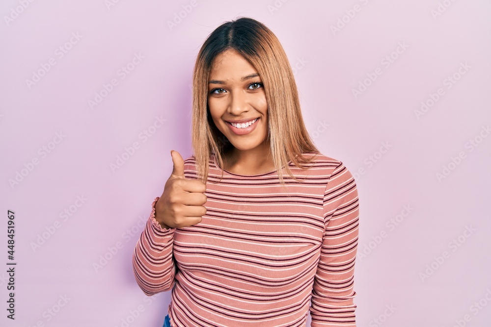 Beautiful hispanic woman wearing casual striped sweater doing happy thumbs up gesture with hand. approving expression looking at the camera showing success. Beautiful hispanic woman wearing casual striped sweater doing happy thumbs up gesture with hand. approving expression looking at the camera showing success.