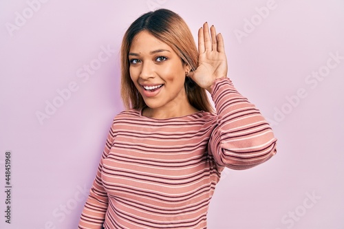 Beautiful hispanic woman wearing casual striped sweater smiling with hand over ear listening an hearing to rumor or gossip. deafness concept.