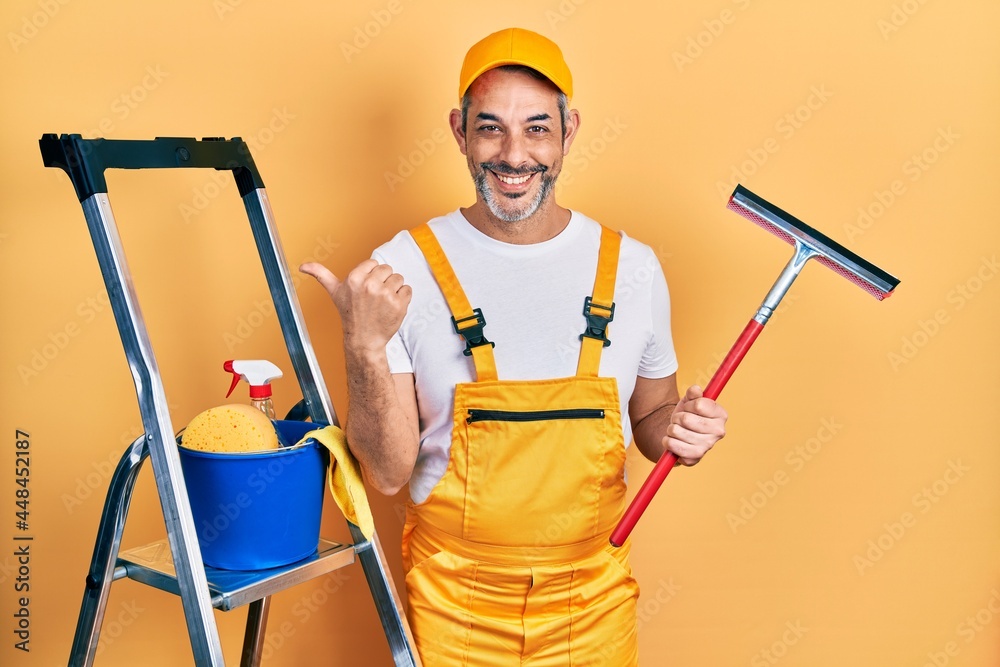 Handsome middle age man with grey hair wearing glass cleaner uniform and squeegee pointing thumb up to the side smiling happy with open mouth Handsome middle age man with grey hair wearing glass cleaner uniform and squeegee pointing thumb up to the side smiling happy with open mouth