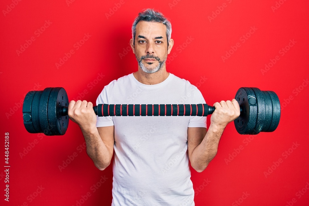 Handsome middle age man with grey hair wearing sportswear using dumbbells relaxed with serious expression on face. simple and natural looking at the camera. Handsome middle age man with grey hair wearing sportswear using dumbbells relaxed with serious expression on face. simple and natural looking at the camera.