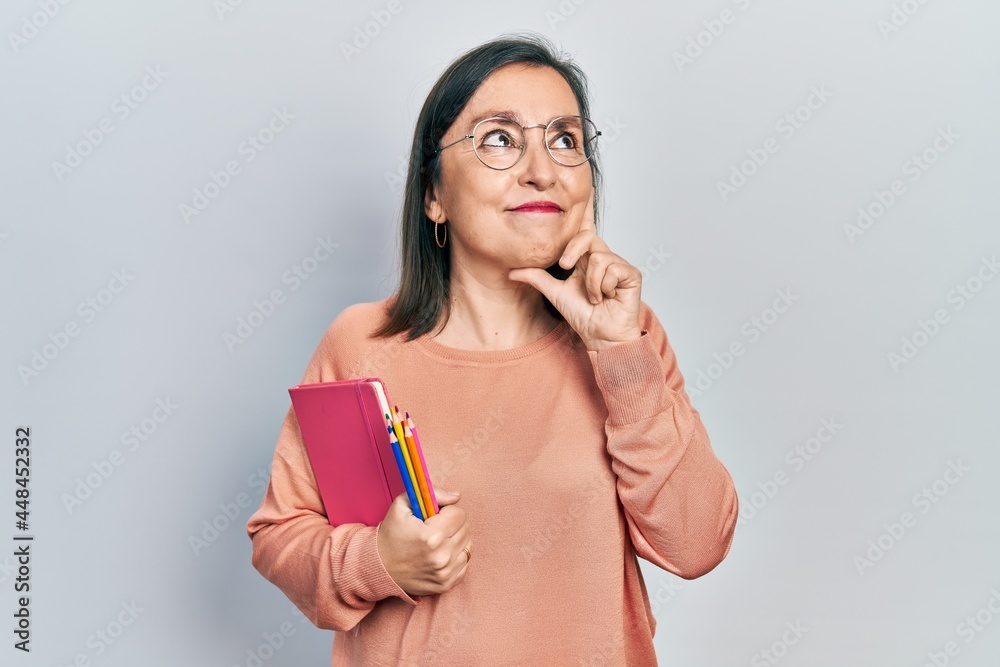 Middle age hispanic woman holding book and color pencils serious face thinking about question with hand on chin, thoughtful about confusing idea