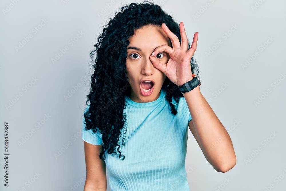 Young hispanic woman with curly hair wearing casual blue t shirt doing ok gesture shocked with surprised face, eye looking through fingers. unbelieving expression. Young hispanic woman with curly hair wearing casual blue t shirt doing ok gesture shocked with surprised face, eye looking through fingers. unbelieving expression.