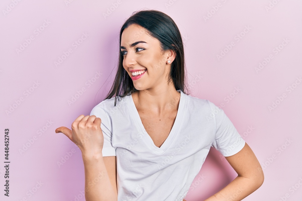 Beautiful woman with blue eyes wearing casual white t shirt pointing thumb up to the side smiling happy with open mouth