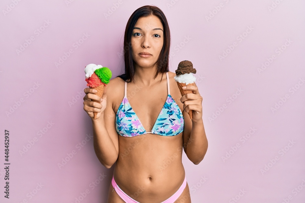 Naklejka premium Young latin woman wearing bikini holding ice cream relaxed with serious expression on face. simple and natural looking at the camera.