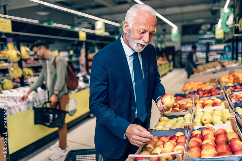 Senior bearded man buying fruits at the supermarket. Other customers in background. Consumerism concept. Senior bearded man buying fruits at the supermarket. Other customers in background. Consumerism concept.