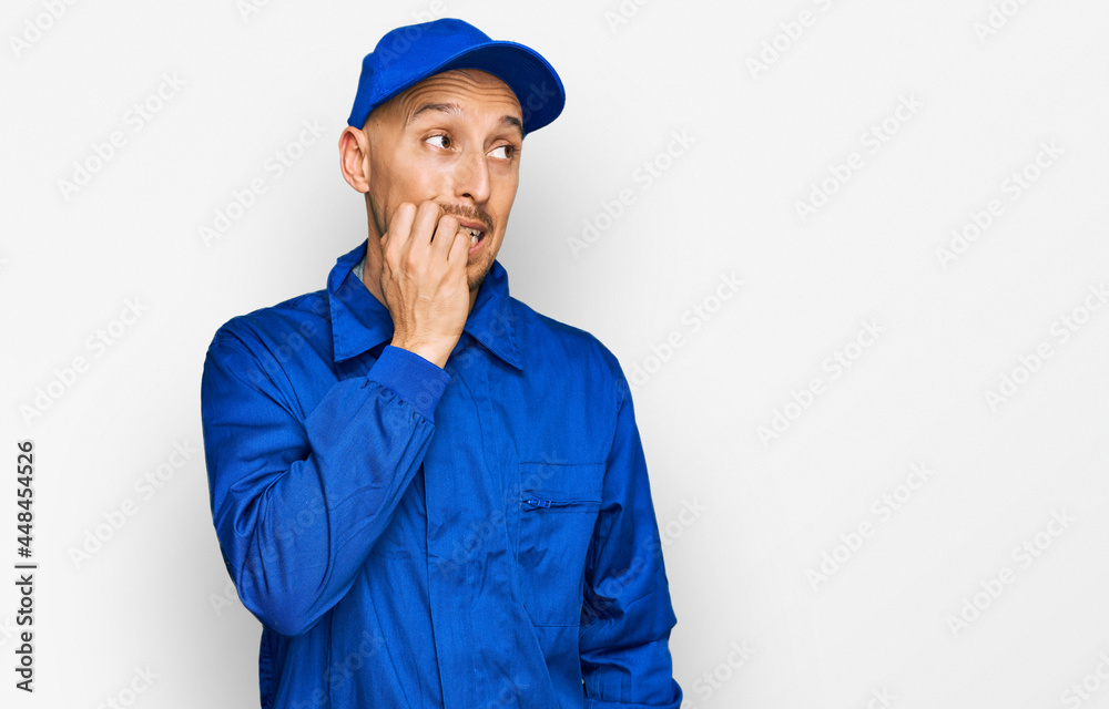 Bald man with beard wearing builder jumpsuit uniform looking stressed and nervous with hands on mouth biting nails. anxiety problem.