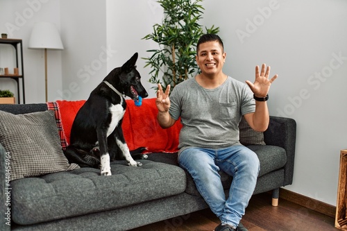 Young latin man and dog sitting on the sofa at home showing and pointing up with fingers number eight while smiling confident and happy. © Krakenimages.com