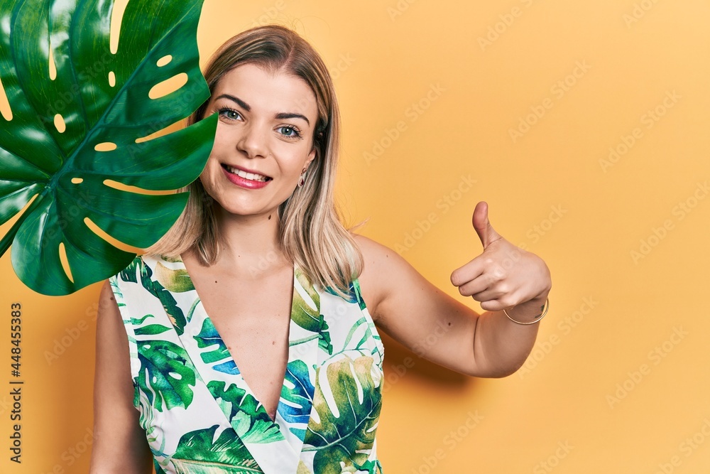 Beautiful caucasian woman with green plant leaf close to beautiful face smiling happy and positive, thumb up doing excellent and approval sign Beautiful caucasian woman with green plant leaf close to beautiful face smiling happy and positive, thumb up doing excellent and approval sign