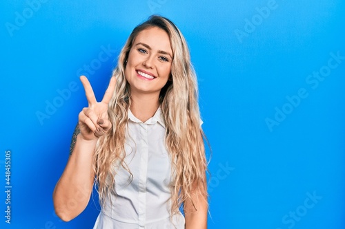 Beautiful young blonde woman wearing casual white shirt showing and pointing up with fingers number two while smiling confident and happy.