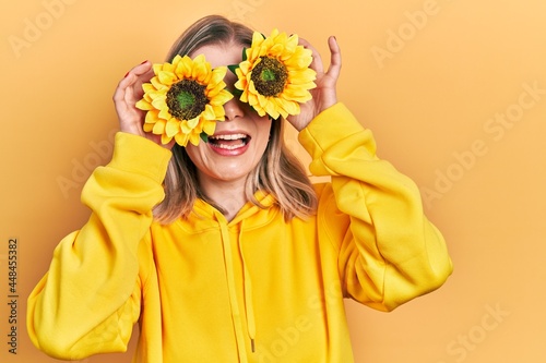 Beautiful caucasian woman holding yellow sunflowers over eyes smiling and laughing hard out loud because funny crazy joke.