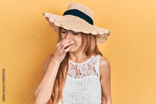 Young caucasian woman wearing summer hat smelling something stinky and disgusting, intolerable smell, holding breath with fingers on nose. bad smell