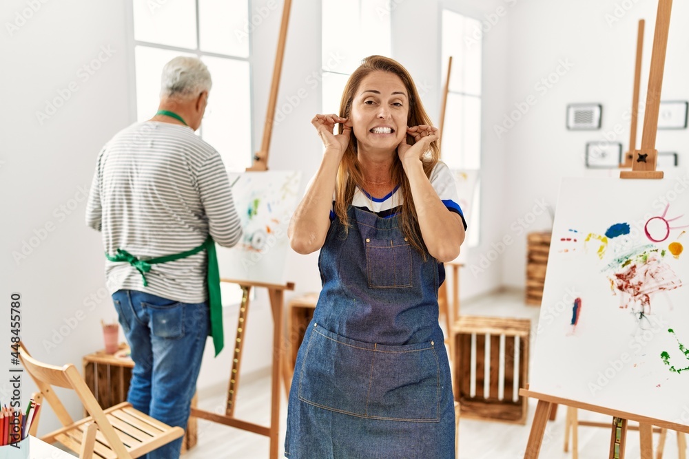 Hispanic woman wearing apron at art studio covering ears with fingers with annoyed expression for the noise of loud music. deaf concept. Hispanic woman wearing apron at art studio covering ears with fingers with annoyed expression for the noise of loud music. deaf concept.