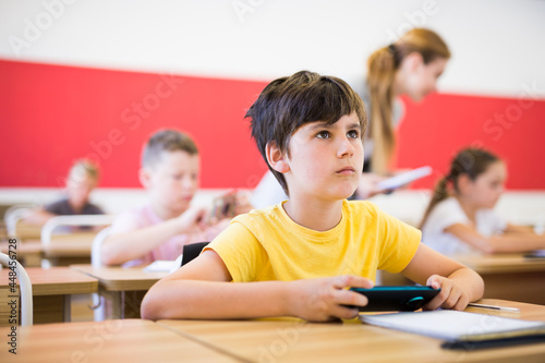 Focused diligent tween schoolboy sitting at lesson in classroom, using mobile phone