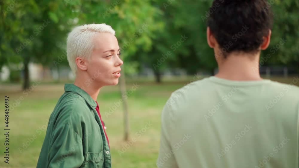 Side view tracking slow motion shot of young couple enjoying walking and talking over coffee in park. Cheerful woman with short blonde hair telling story to boyfriend and laughing Side view tracking slow motion shot of young couple enjoying walking and talking over coffee in park. Cheerful woman with short blonde hair telling story to boyfriend and laughing