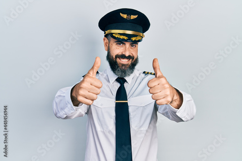 Young hispanic man wearing airplane pilot uniform approving doing positive gesture with hand, thumbs up smiling and happy for success. winner gesture.