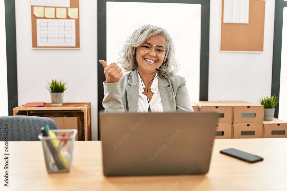 Middle age businesswoman sitting on desk working using laptop at office smiling with happy face looking and pointing to the side with thumb up. Middle age businesswoman sitting on desk working using laptop at office smiling with happy face looking and pointing to the side with thumb up.