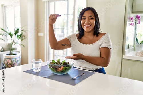 Young hispanic woman eating healthy salad at home gesturing with hands showing big and large size sign, measure symbol. smiling looking at the camera. measuring concept.