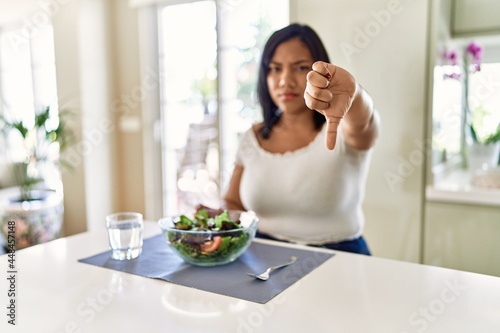 Young hispanic woman eating healthy salad at home looking unhappy and angry showing rejection and negative with thumbs down gesture. bad expression.