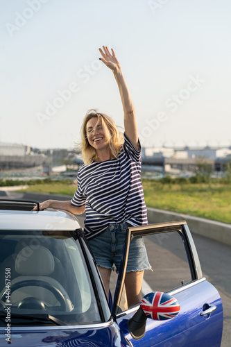Excited woman standing at driver seat of new car wave hand. Cheerful female of middle age happy of buying automobile in dealership or of renting vehicle for road trip vacation. Auto transport concept © DimaBerlin