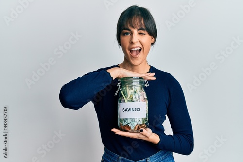 Young hispanic woman holding savings jar winking looking at the camera with sexy expression, cheerful and happy face.