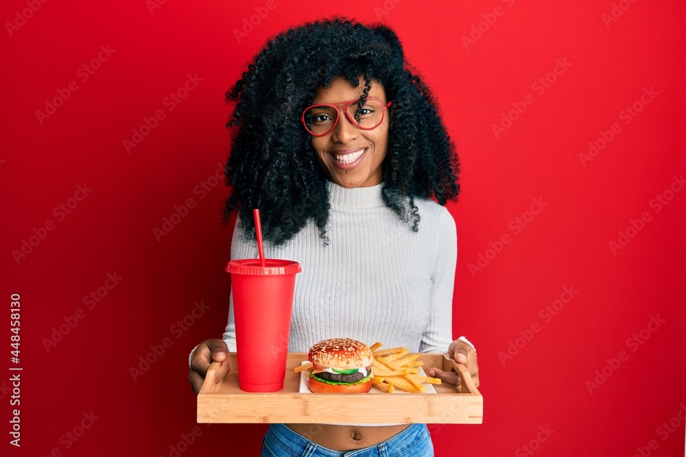 African american woman with afro hair eating a tasty classic burger with fries and soda smiling with a happy and cool smile on face. showing teeth. African american woman with afro hair eating a tasty classic burger with fries and soda smiling with a happy and cool smile on face. showing teeth.