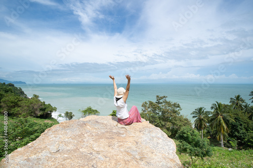 Happy young asian woman with hands up on the cliff watching  the sea on sunny day. A female in casual dress on ocean cliff having fun listening to the waves and breathing sea breeze. photo