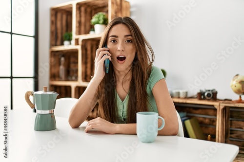 Young hispanic woman talking on the smartphone sitting on the table at home scared and amazed with open mouth for surprise, disbelief face