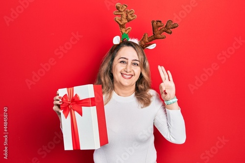 Middle age caucasian woman wearing cute christmas reindeer horns holding gift doing ok sign with fingers, smiling friendly gesturing excellent symbol
