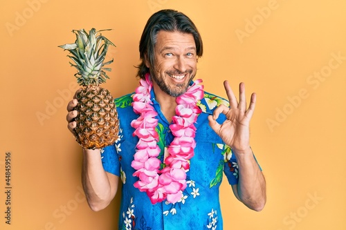 Middle age handsome man wearing hawaiian lei holding pineapple doing ok sign with fingers, smiling friendly gesturing excellent symbol