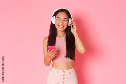 Portrait of carefree attractive asian woman enjoying favorite song, close eyes to relax while listening music in hearphones, holding mobile phone and standing over pink background photo