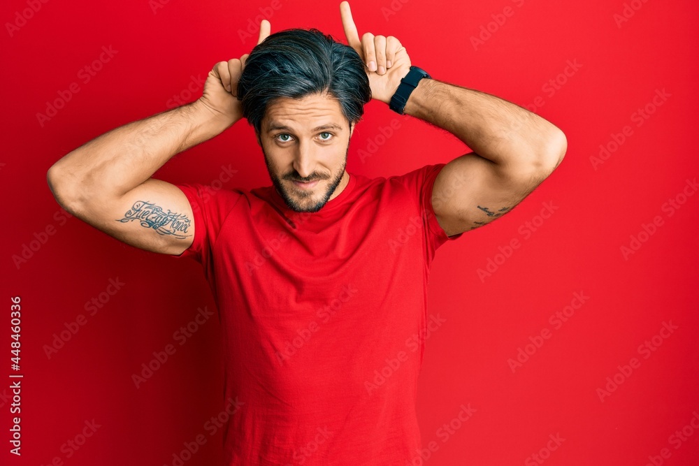 Young hispanic man wearing casual red t shirt doing funny gesture with finger over head as bull horns Young hispanic man wearing casual red t shirt doing funny gesture with finger over head as bull horns