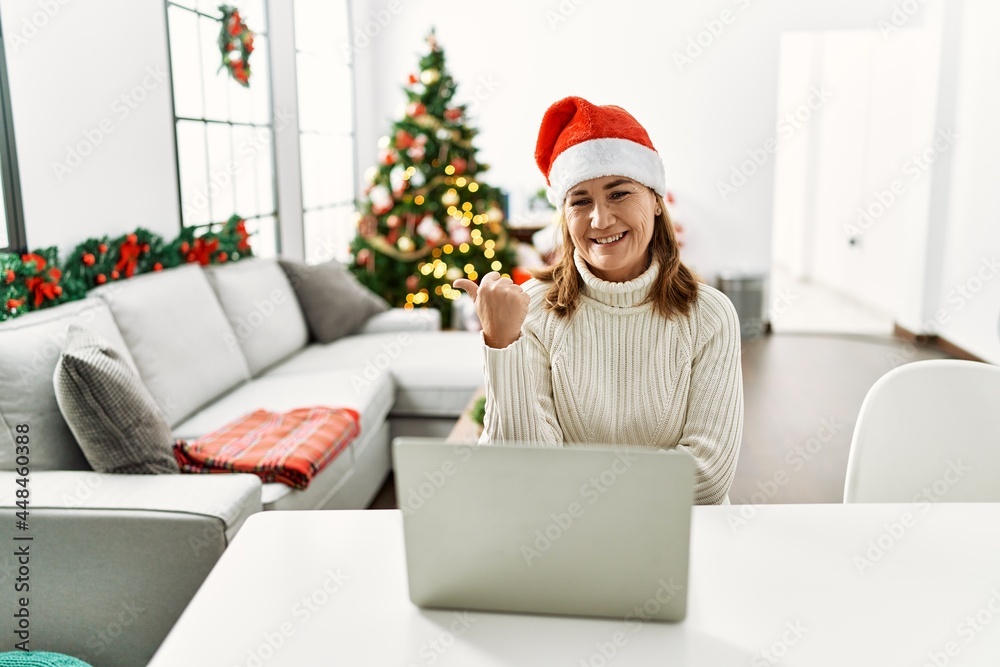 Middle age woman wearing santa claus hat using laptop smiling with happy face looking and pointing to the side with thumb up. Middle age woman wearing santa claus hat using laptop smiling with happy face looking and pointing to the side with thumb up.