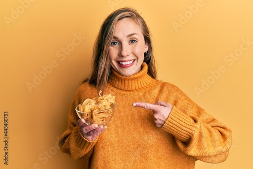 Young blonde woman holding bowl with uncooked pasta smiling happy pointing with hand and finger