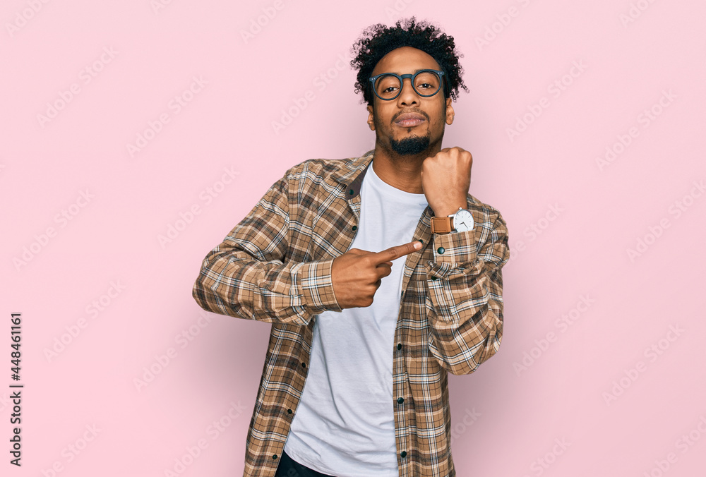 Young african american man with beard wearing casual clothes and glasses in hurry pointing to watch time, impatience, looking at the camera with relaxed expression Young african american man with beard wearing casual clothes and glasses in hurry pointing to watch time, impatience, looking at the camera with relaxed expression