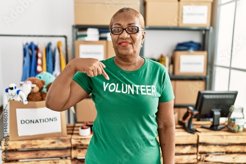 Mature hispanic woman wearing volunteer t shirt at donations stand smiling happy pointing with hand and finger © Krakenimages.com