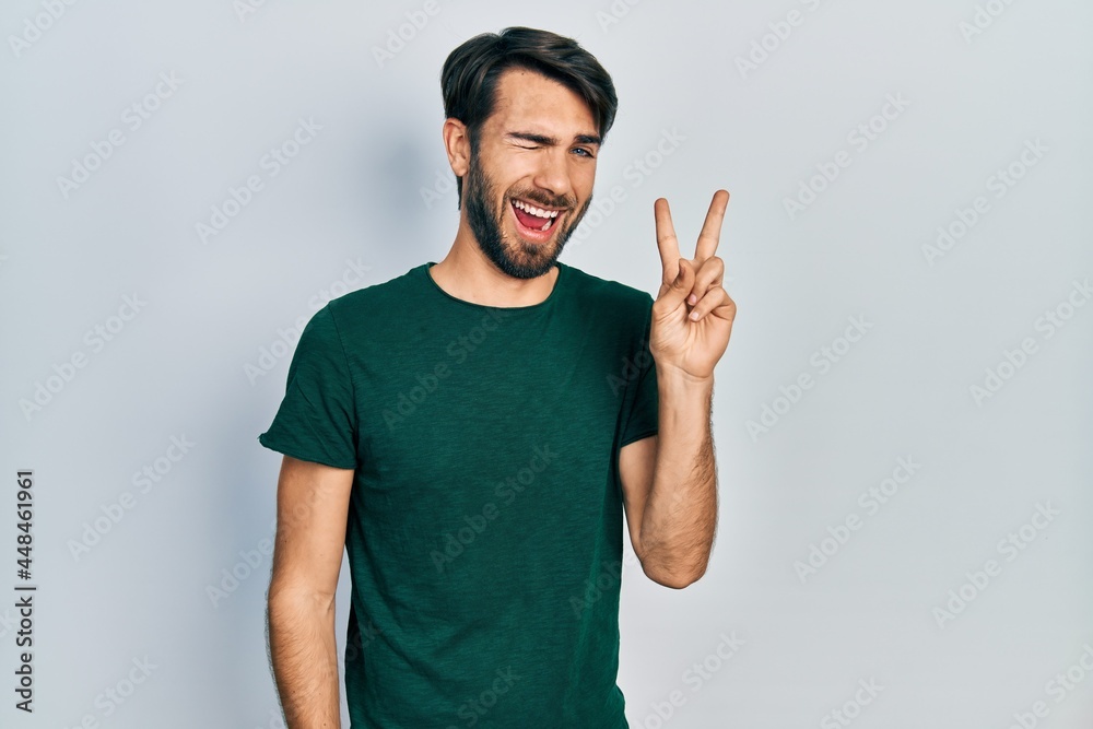 Fototapeta premium Young hispanic man wearing casual white tshirt smiling with happy face winking at the camera doing victory sign. number two.
