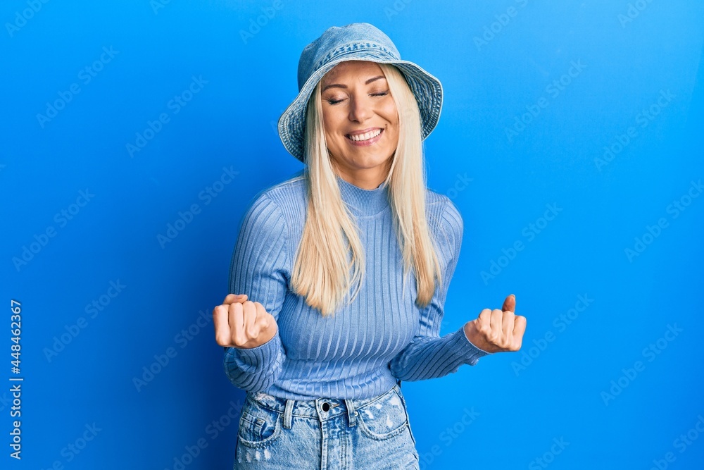 Young blonde woman wearing casual denim hat very happy and excited doing winner gesture with arms raised, smiling and screaming for success. celebration concept. Young blonde woman wearing casual denim hat very happy and excited doing winner gesture with arms raised, smiling and screaming for success. celebration concept.