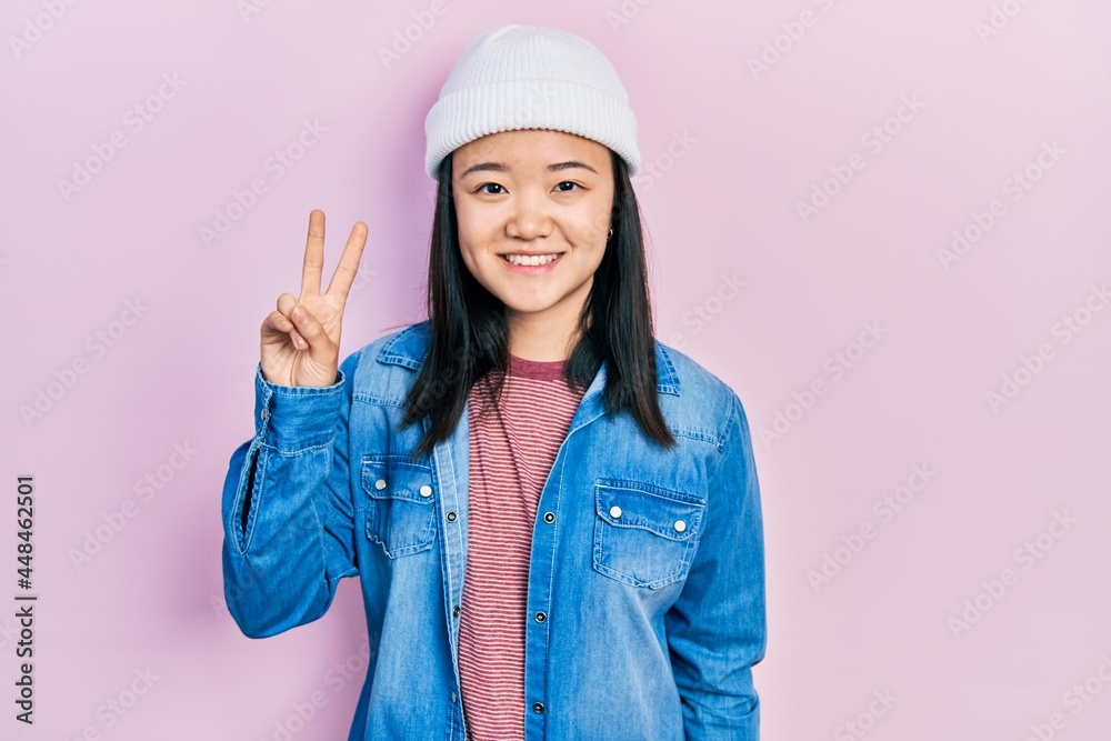Young chinese girl wearing cute wool cap smiling with happy face winking at the camera doing victory sign. number two. Young chinese girl wearing cute wool cap smiling with happy face winking at the camera doing victory sign. number two.