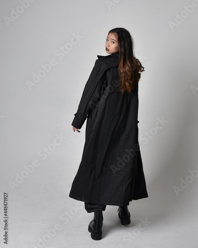 Full length portrait of pretty young asian girl wearing black tank top, utilitarian  pants, leather boots and large dark coat. Standing pose with gestural hand movements,  isolated on  studio backgrou