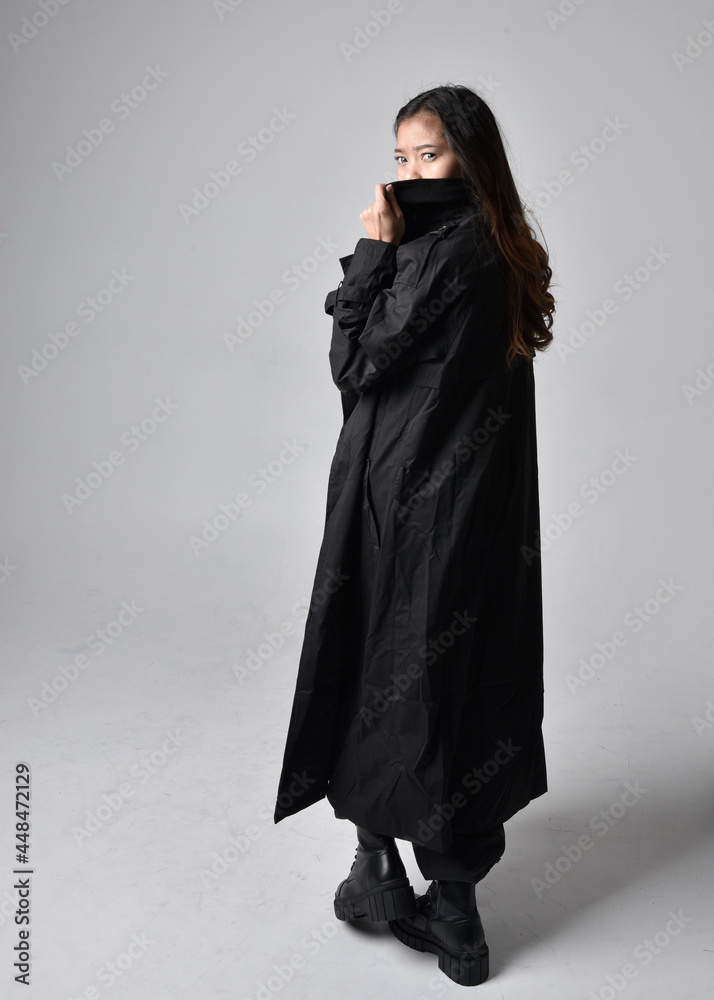 Full length portrait of pretty young asian girl wearing black tank top, utilitarian pants, leather boots and large dark coat. Standing pose with gestural hand movements, isolated on studio backgrou Full length portrait of pretty young asian girl wearing black tank top, utilitarian pants, leather boots and large dark coat. Standing pose with gestural hand movements, isolated on studio backgrou