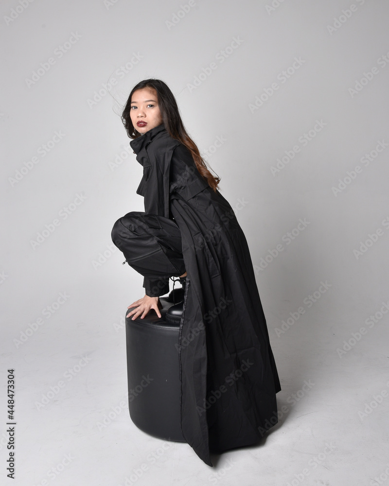 Full length portrait of pretty young asian girl wearing black tank top, utilitarian pants, leather boots and large dark coat. Sitting pose with gestural hand movements, isolated on studio backgroun Full length portrait of pretty young asian girl wearing black tank top, utilitarian pants, leather boots and large dark coat. Sitting pose with gestural hand movements, isolated on studio backgroun