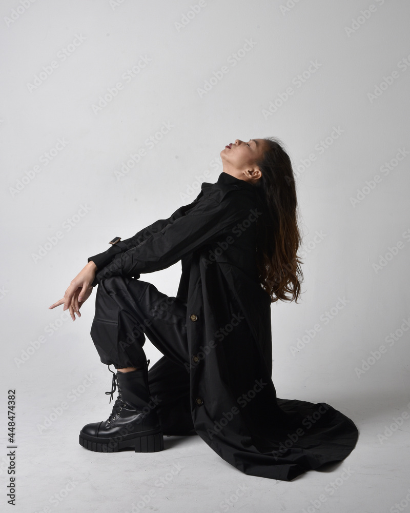 Full length portrait of pretty young asian girl wearing black tank top, utilitarian  pants, leather boots and large dark coat. Sitting pose with gestural hand movements,  isolated on  studio backgroun