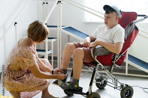 Mother helping disabled boy 17 years old in wheelchair with cerebral palsy to put on shoes, before going for a walk. Woman assists person with Special Need in rehabilitation canter. Inclusion, access photo