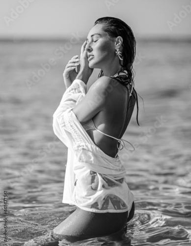 Sexy woman girl in white bikini and shirt stands sideways to camera in waist-deep clear sea water, facing sun during vacation on the seashore. Black and white. Summer vibes. On the beach